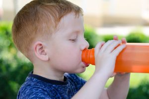Young boy drinking from an orange water bottle outdoors.